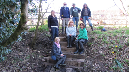 A group of people wearing coats sit on a set of stone steps surrounded by trees and smile at the camera