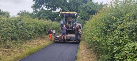 A construction vehicle surrounded by people sits on a newly resurfaced road, with hedgerows on each side