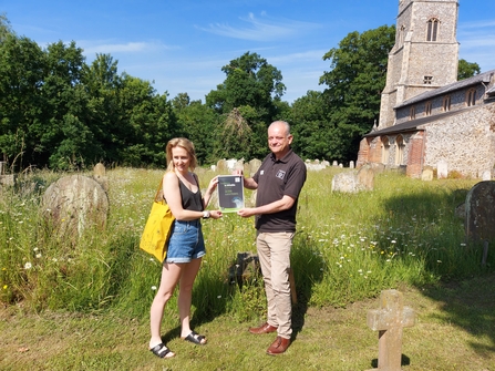 Two people in tshirts stand holding a certificate in a churchyard on a sunny day