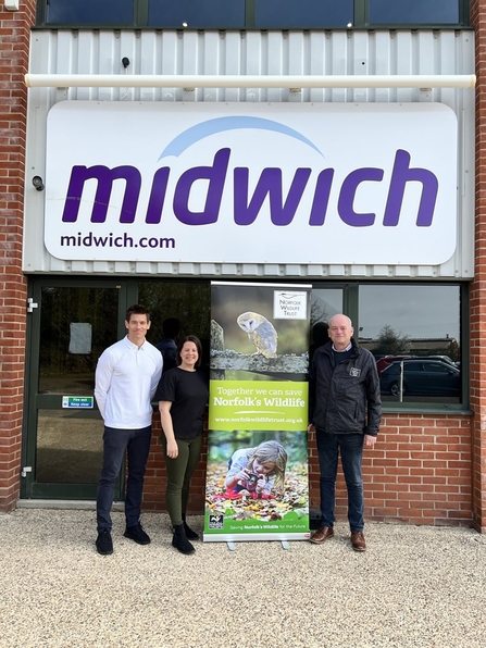 Three people stand either side of a Norfolk Wildlife Trust banner in front of a building with a Midwich logo attached to the wall
