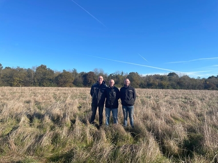 Three men stand in a line in a field, smiling at the camera, under a bright blue sky on a sunny day