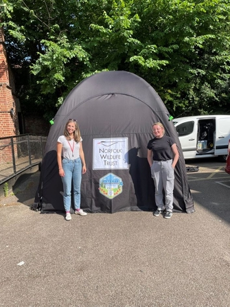 Two people stand in front of a black tent with the Norfolk Wildlife Trust logo on the front
