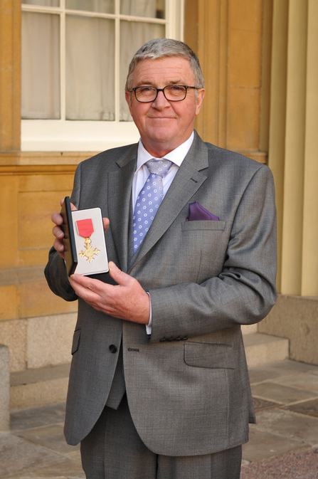 A man wearing a grey suit and purple tie smiles at the camera while holding a medal