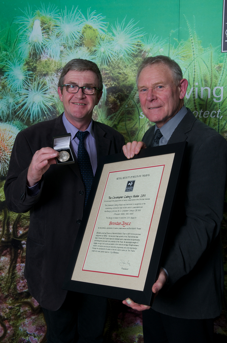 Two men stand beside each other smiling at the camera, with one holding a medal and the other holding a poster