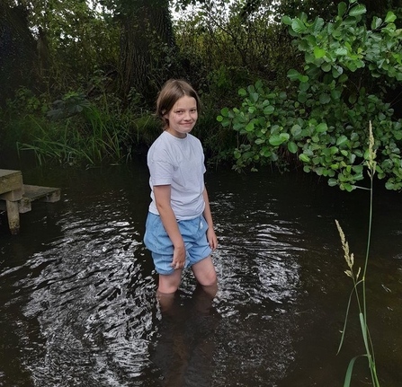 A young person with short brown hair smiles as they stand knee deep in a river