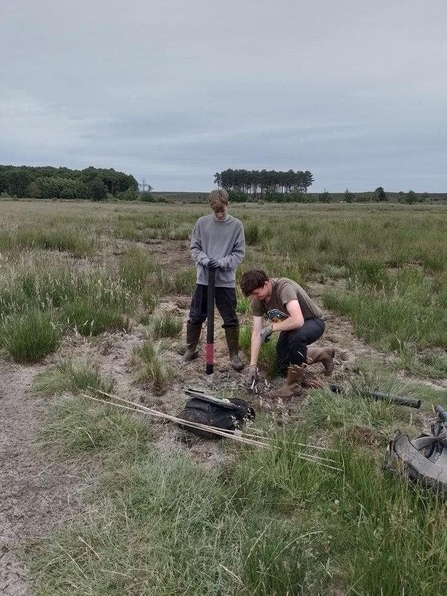 Two students install ground water monitoring tubes in the ground in a field on a cloudy day