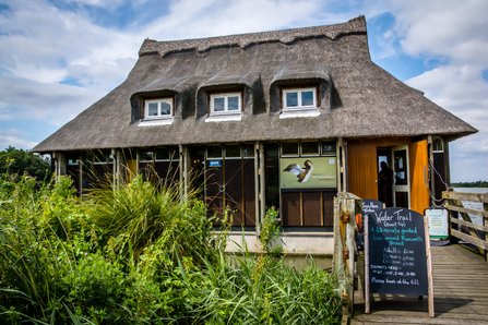 Ranworth Broad thatched visitor centre in the summer sun, with lush green vegetation in front and a boat trip timetable