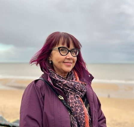 Mandy Loadman is smiling on a beach. She has purple hair and a purple coat and scarf and is wearing glasses.