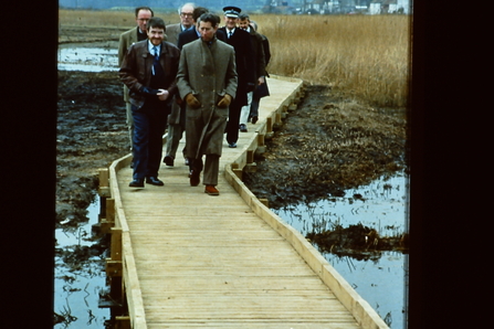 Prince Charles walks along a boardwalk, leading a group of people, as they look across the marshes