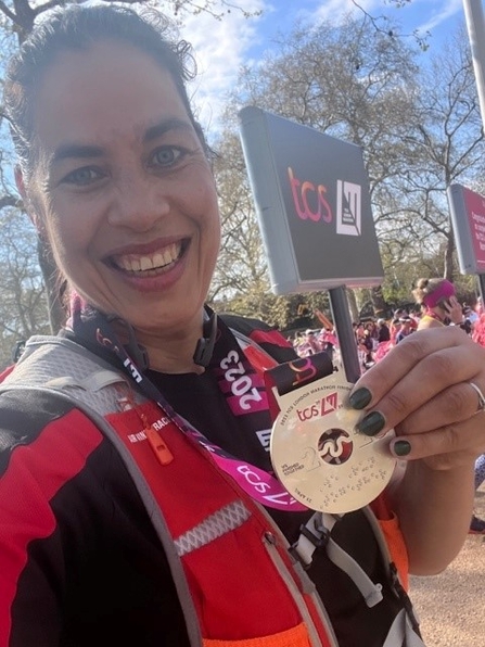 A woman wearing sports clothes holds a medal to the camera and smiles, in front of a crowd of runners