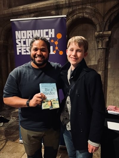 A man with black hair and a beard holds a book called 'Be a Birder'. He stands beside a teenage boy with blond hair. They are both smiling at the camera, in front of a poster for Norwich Science Festival.