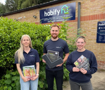 Two women and a man stand in front of an industrial building. They are holding Norfolk Wildlife Trust leaflets and an Investors in Wildlife certificate.