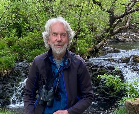 David North posing with his binoculars in front of a small waterfall. He has wavy grey hair and a grey beard.