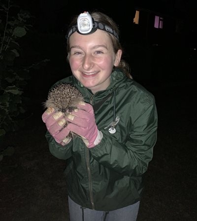 Poppy is grinning at night with a head torch on holding a hedgehog. She is wearing pink gloves. 