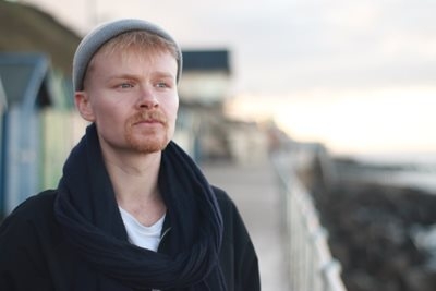 James is posing in a beanie with beach huts in the background.