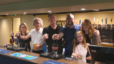 Three men, two women and a young girl stand behind the bar of a pub, smiling at the camera, while some of them pretend to pull a pint