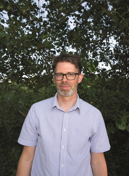 Norfolk Wildlife Trust CEO, Eliot Lyne, wearing a light blue shirt and standing in front of a tree