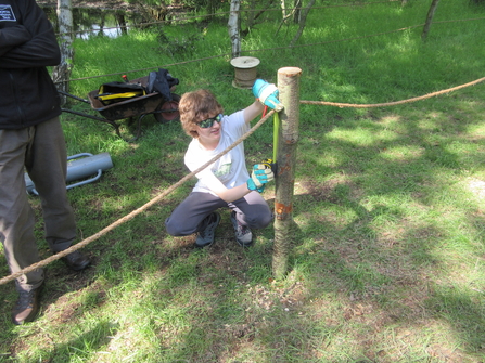 Erin is measuring a wooden post for a rope and wood fence he is making. 