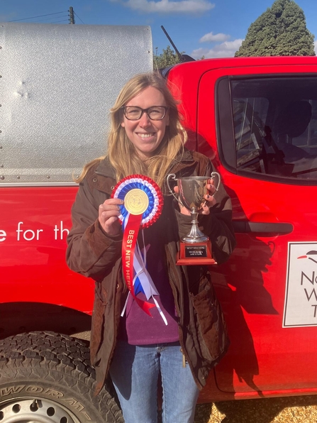 Laura is smiling and holding up a rosette and cup for best new heard. She is standing in front of a red NWT vehicle. 