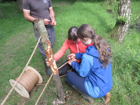 Two young people with long brown hair and wearing waterproof coats are holding a tree stump and some rope, as they work to build a fence