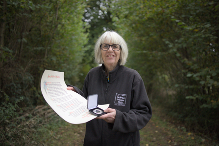 A woman with short grey hair wearing glasses smiles at the camera while holding a medal in a case and a large piece of paper. There are hedges and green grass in the background.