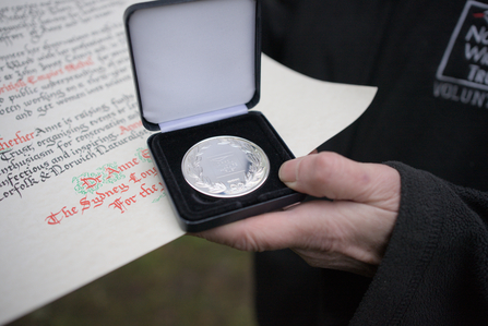 A person's hand holding a silver medal inside a case, with a scroll of paper behind it featuring cursive text.