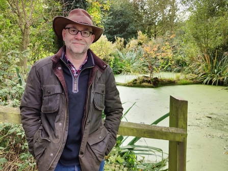 Richard is wearing a big brown hat and is smiling in front of a green body of water with lots of trees and shrubs around. 