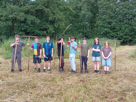 A group of young people stand in a line in a field, all looking at the camera. Many of them are holding large rakes.