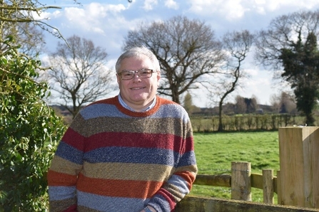 Richard has short grey hair and glasses, he is wearing a colourful stripey jumper and leaning against a wooden fence with trees in the background. 