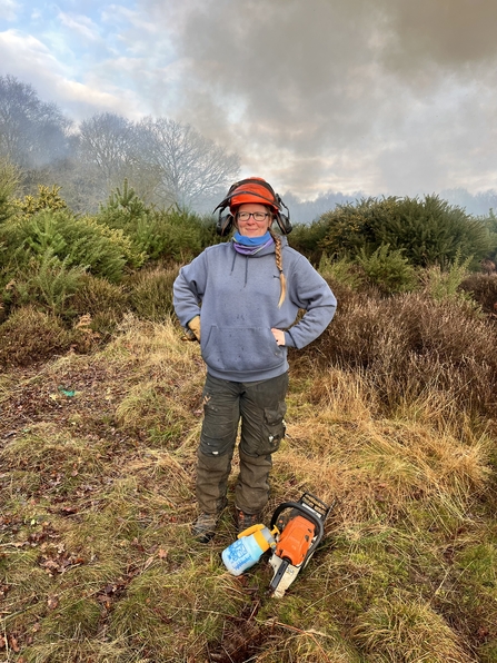 Anne is posing outdoors with her hands on her hips. She has a hard hat on and there are tools at her feet.