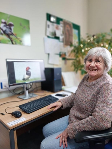 Our volunteer, Carol, grinning at her desk at Bewick House. She has grey hair and glasses and is wearing a mauve coloured jumper and blue jeans. There is a photo of a duck on her computer screen. 