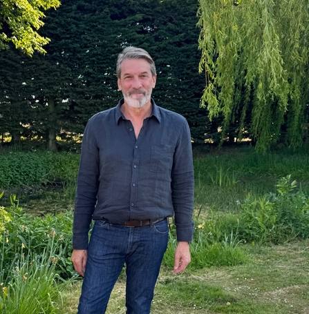 A smiling, tall man with grey hair and a grey beard standing outside on a sunny day beside a willow tree. He is wearing a navy shirt and dark blue jeans.