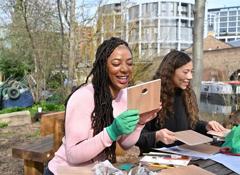 Two young people sit together smiling and building bird boxes in a garden by a canal. The person on the left has long black hair in braids and is wearing a pink jumper and green gloves. The person on the right has long curly dark brown hair and is wearing a black jumper.