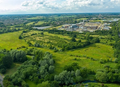 An aerial view over Sweet Briar Marshes. There is a large green space, broken up by patches of green trees and bushes, with houses and business buildings in the distance, all under a blue sky with some fluffy white clouds.