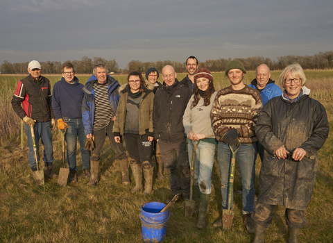 A group of volunteers standing together and smiling at the camera on a sunny winter afternoon. They are wearing wellies and warm clothes, with many holding spades.