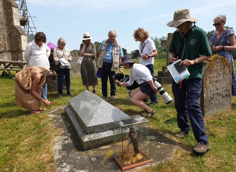 A group of people stand around a gravestone at Wymondham Abbey, looking down at the ground. There is a glass box containing a stuffed bird beside the gravestone.