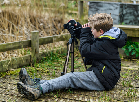 A blond-haired boy wearing a black coat and grey trousers sits on the ground looking through a camera on a tripod