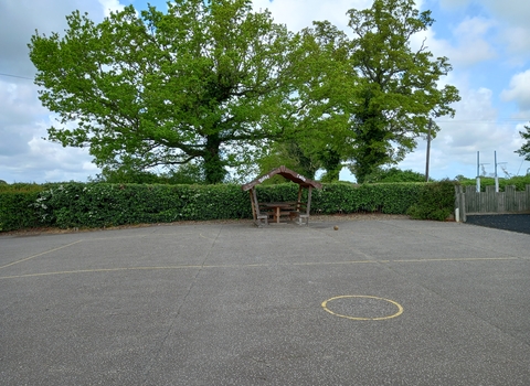 A school playground with a sheltered bench at the back, with trees behind the playground hedge