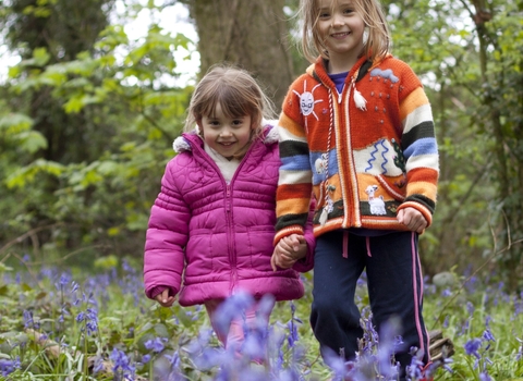 Two children are smiling and standing in a woodland with bluebells