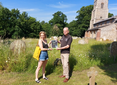 Two people in tshirts stand holding a certificate in a churchyard on a sunny day