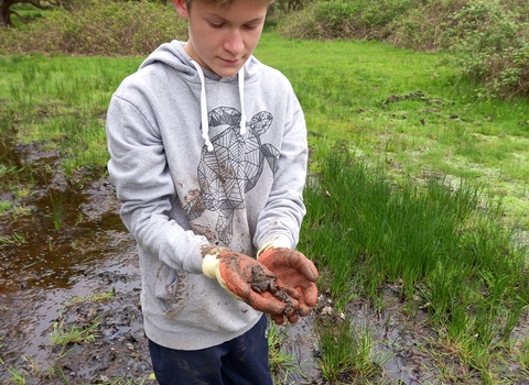 A young person in a grey jumper and black cap holds a toad in his hands
