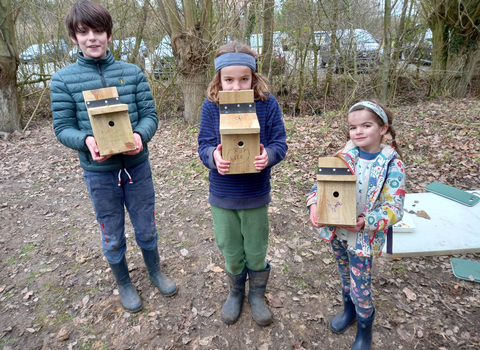 Three children stand in a row in a wooded area holding wooden bird boxes and smiling
