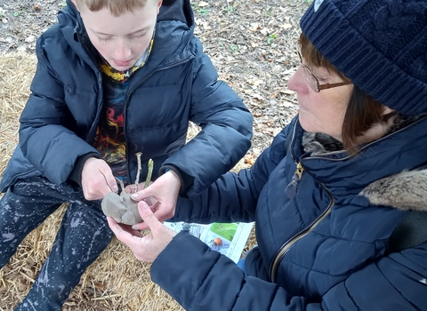 A young boy and a woman wearing winter coats create a spider using clay and sticks