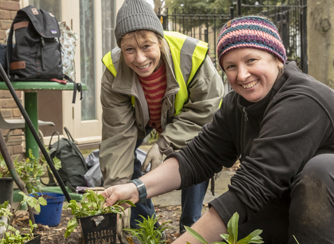 Two smiley community members are watering and potting small plants with big smiles on their faces.