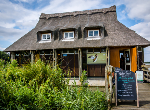 Ranworth Broad thatched visitor centre in the summer sun, with lush green vegetation in front and a boat trip timetable