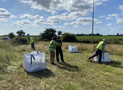 Four people wearing high-vis jackets collecting hay into large bags in a field on a sunny day