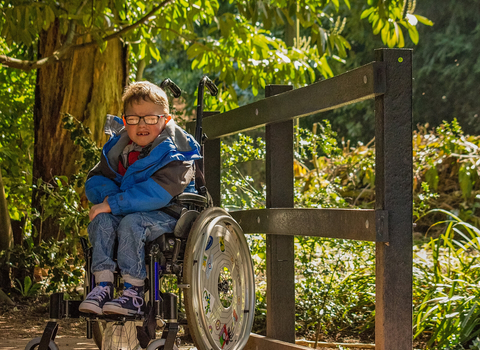 A child wearing a blue coat, jeans and glasses sits in a wheelchair, looking at the camera. They are on a boardwalk, with green trees and bushes behind them on a sunny day